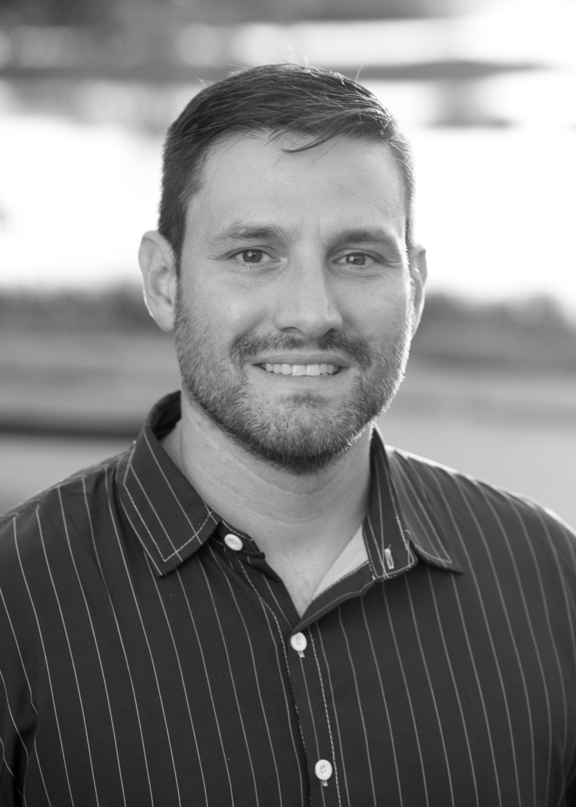 headshot of Aaron Friesz in black and white. White man, dark hair beard and mustache wearing a dark buttoned shirt with light narrow vertical stripes